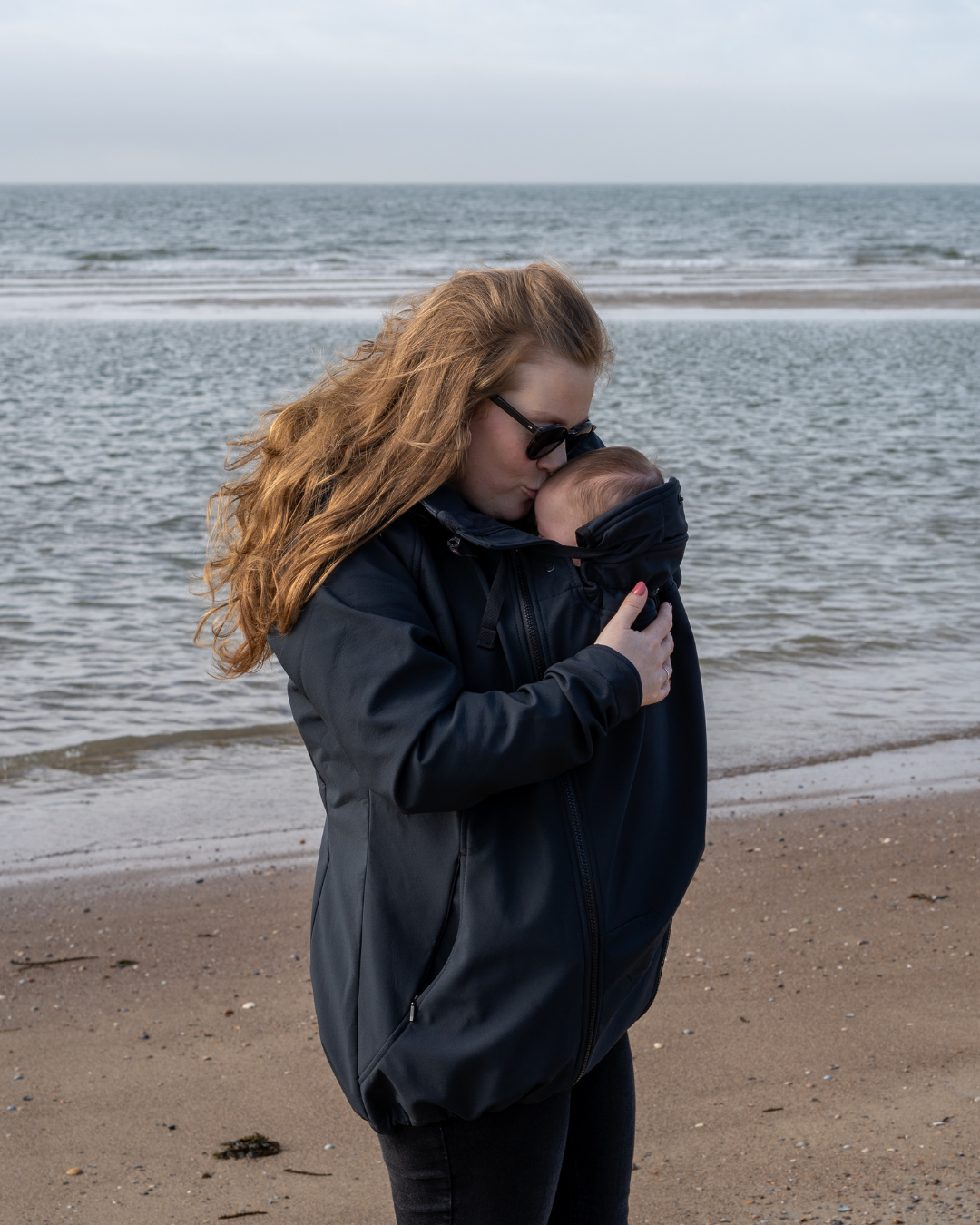 Femme à la plage portant bébé sous veste sombre, embrasse sa tête