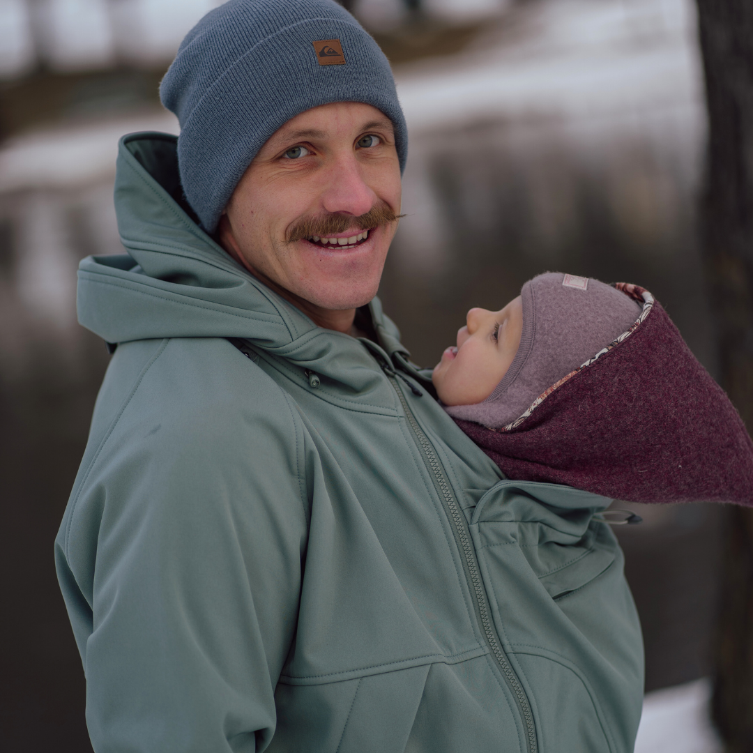 Père portant bébé en veste de portage softshell verte, bien habillés en extérieur