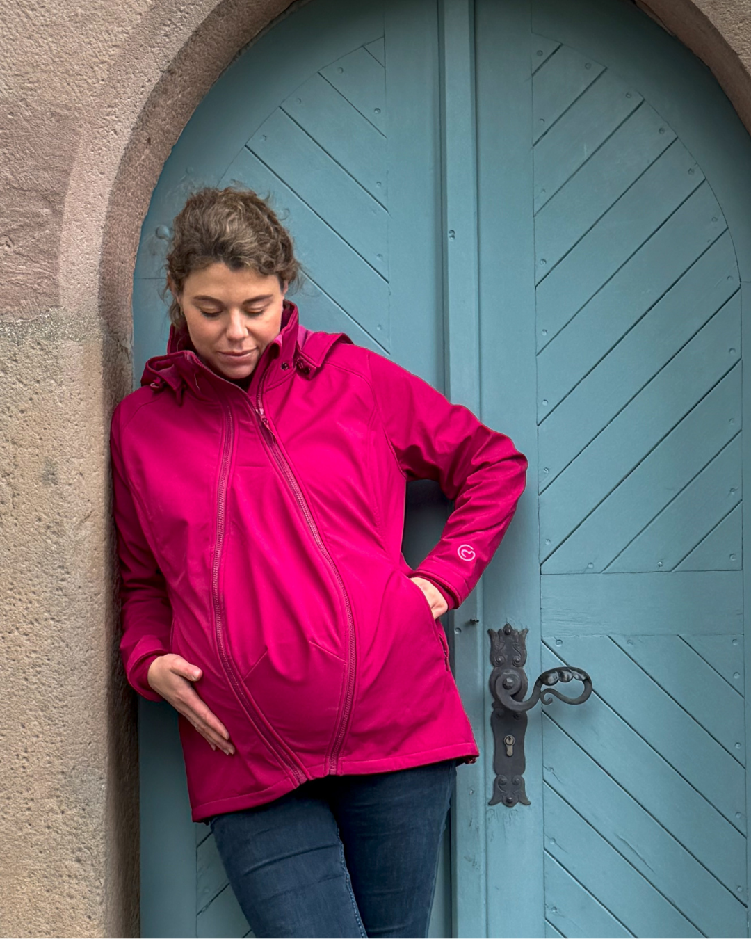 Femme enceinte en veste softshell rose appuyée contre porte bleue, main sur ventre