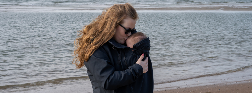 Maman porte bébé en veste de portage sur la plage, mer et vent