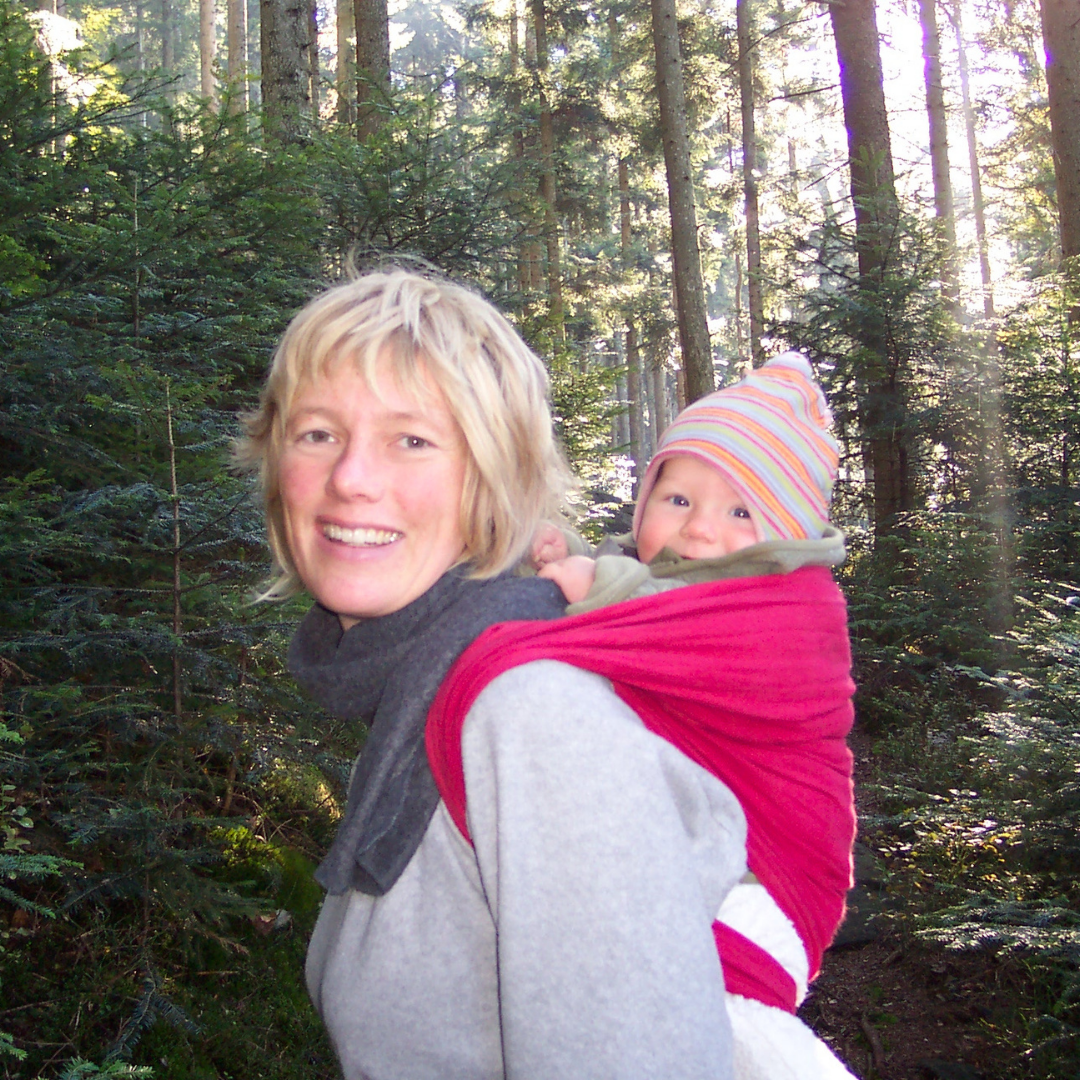 Femme portant bébé dans écharpe rouge au dos, forêt ensoleillée