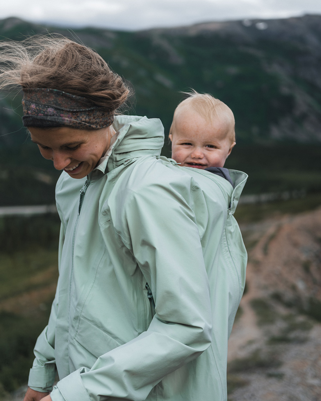 Maman portant bébé sur le dos en veste de portage verte, en randonnée en montagne