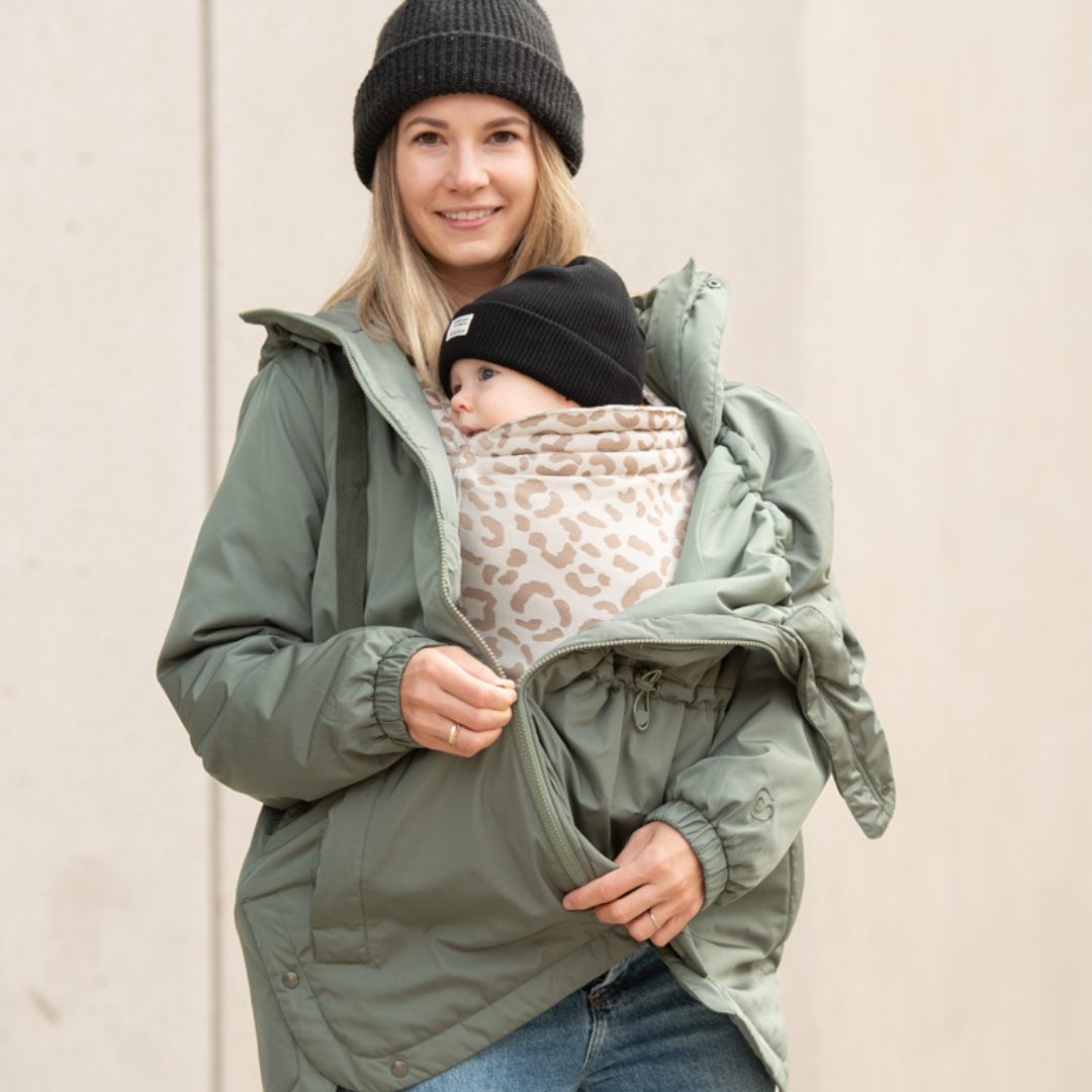 Maman portant bébé en veste de portage verte avec insert à motif, fond urbain