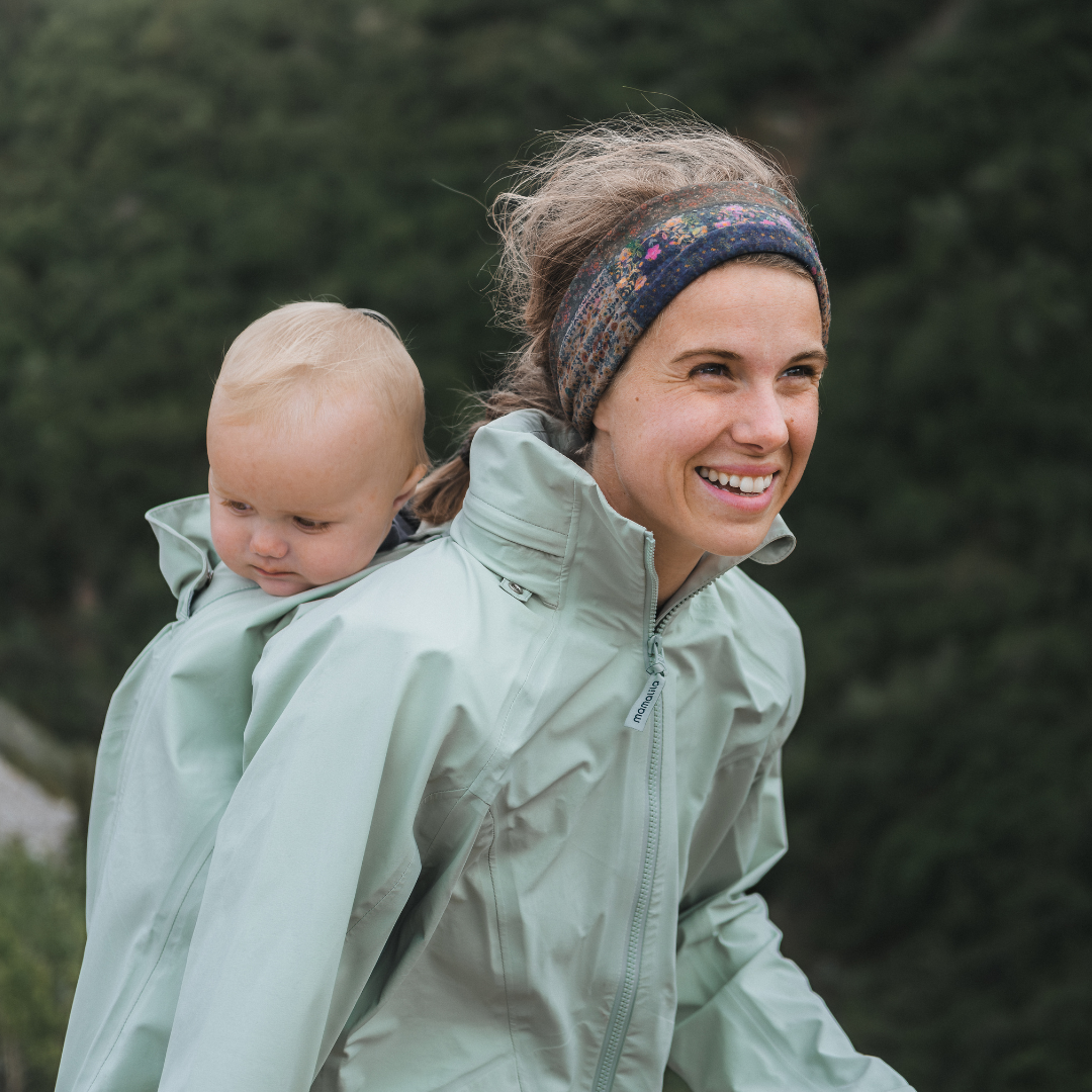 Femme portant bébé au dos avec veste claire, souriante en pleine nature