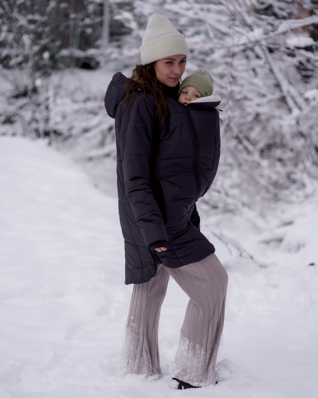Maman portant bébé dans une veste matelassée sombre dans la neige, paysage hivernal
