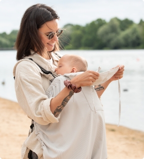 Maman au bord du lac portant bébé avec une couverture de portage claire, lisant un livre